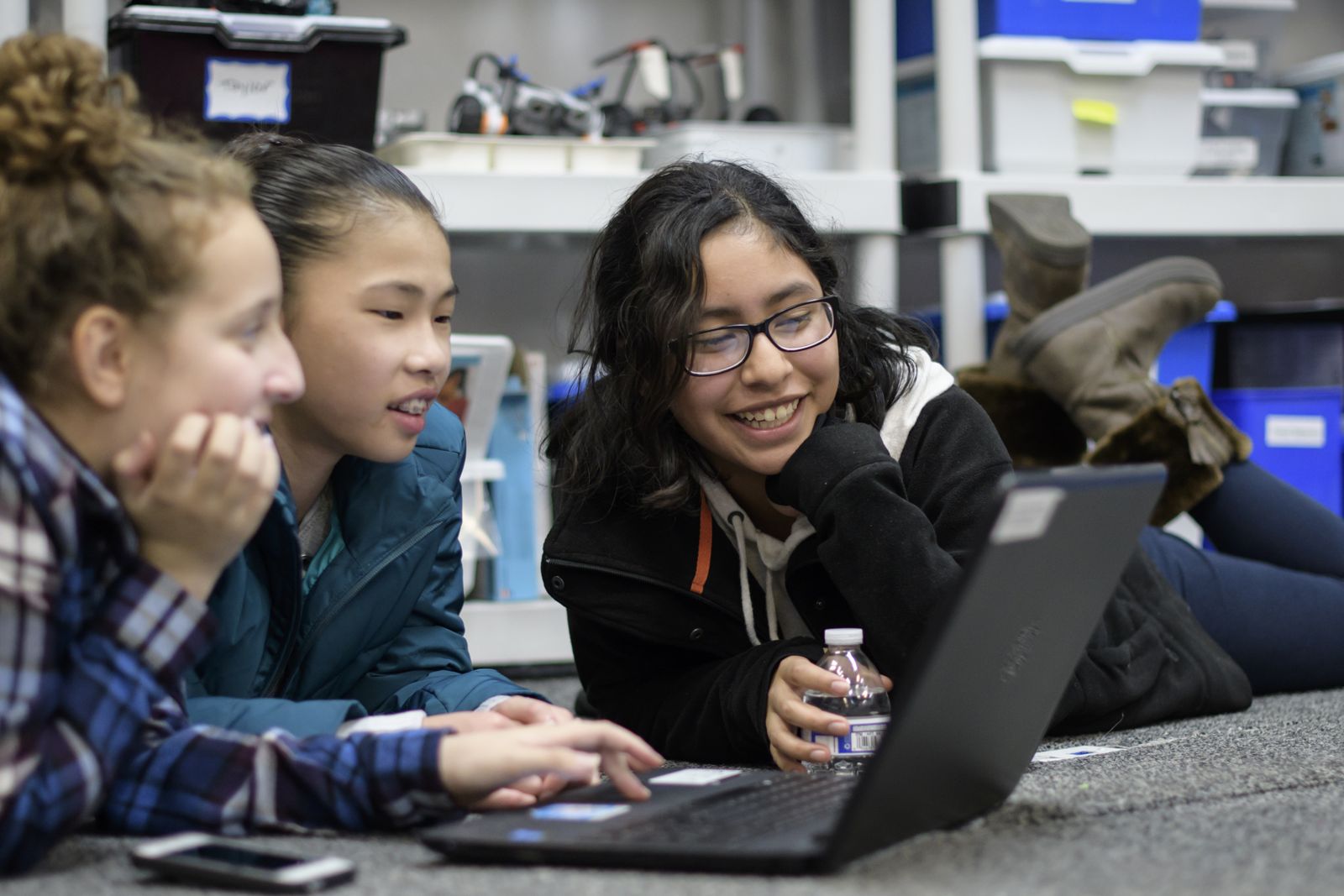 Three students lying on the floor around a laptop, laughing as they code